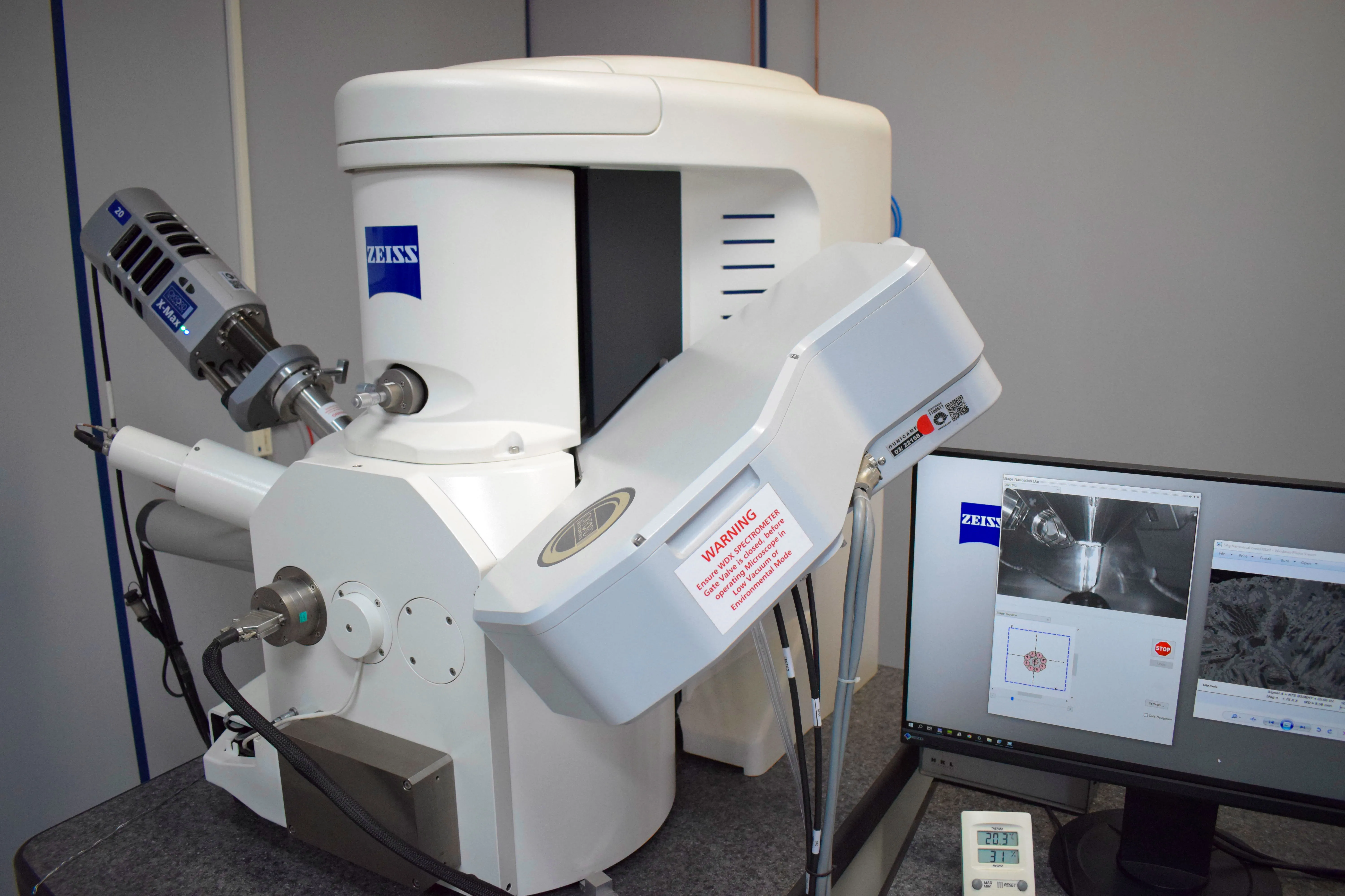 Microscópio eletrônico de varredura (MEV) da marca Zeiss, equipado com detector de raios-X EDS X-Max, acompanhado de um monitor exibindo imagens microscópicas em escala de cinza, com aviso de segurança afixado ao equipamento e termômetro digital posicionado à frente, em ambiente laboratorial.